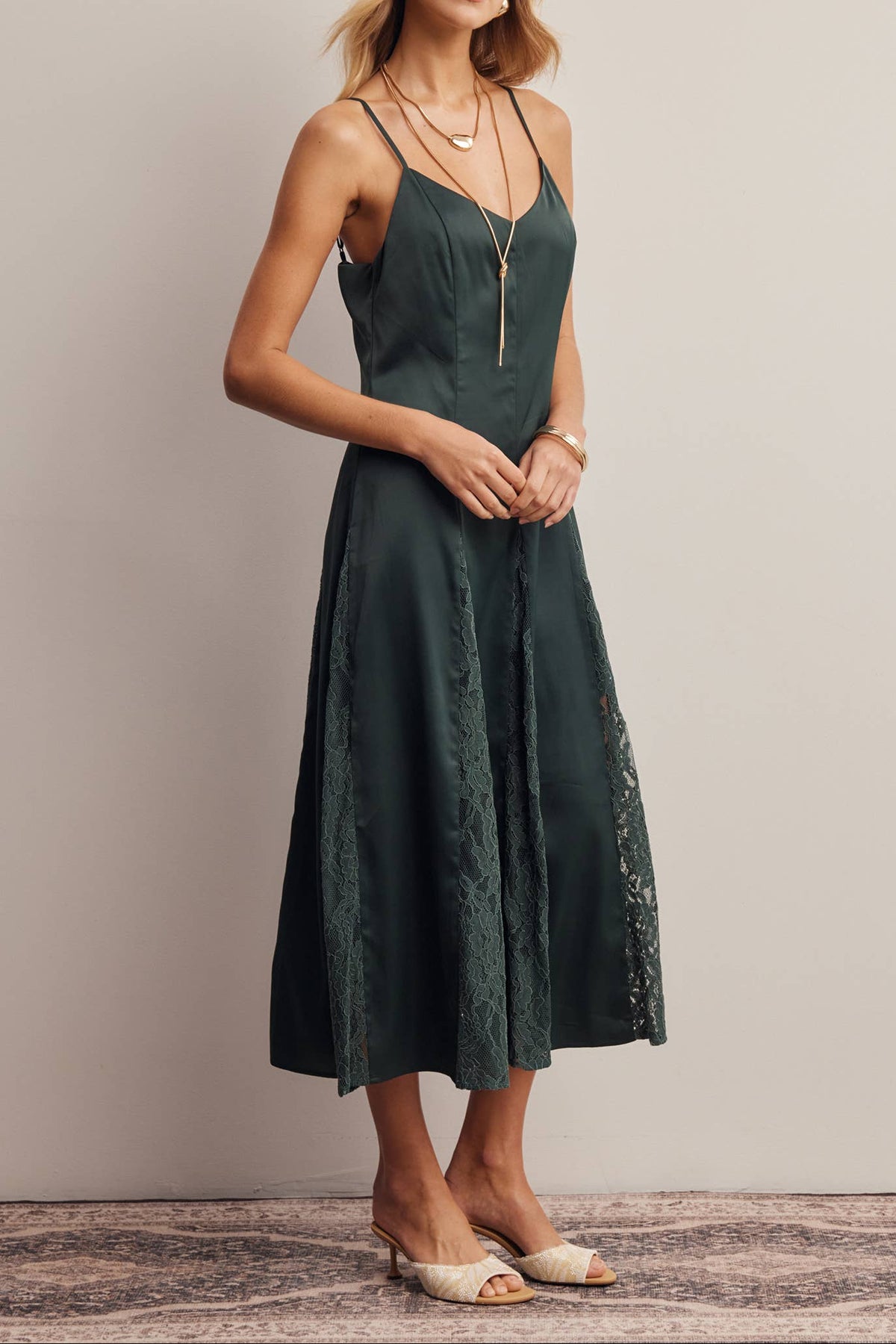 A woman modeling a dark green midi dress with thin straps, a V-neckline, and lace detailing along the sides, standing against a plain beige wall.