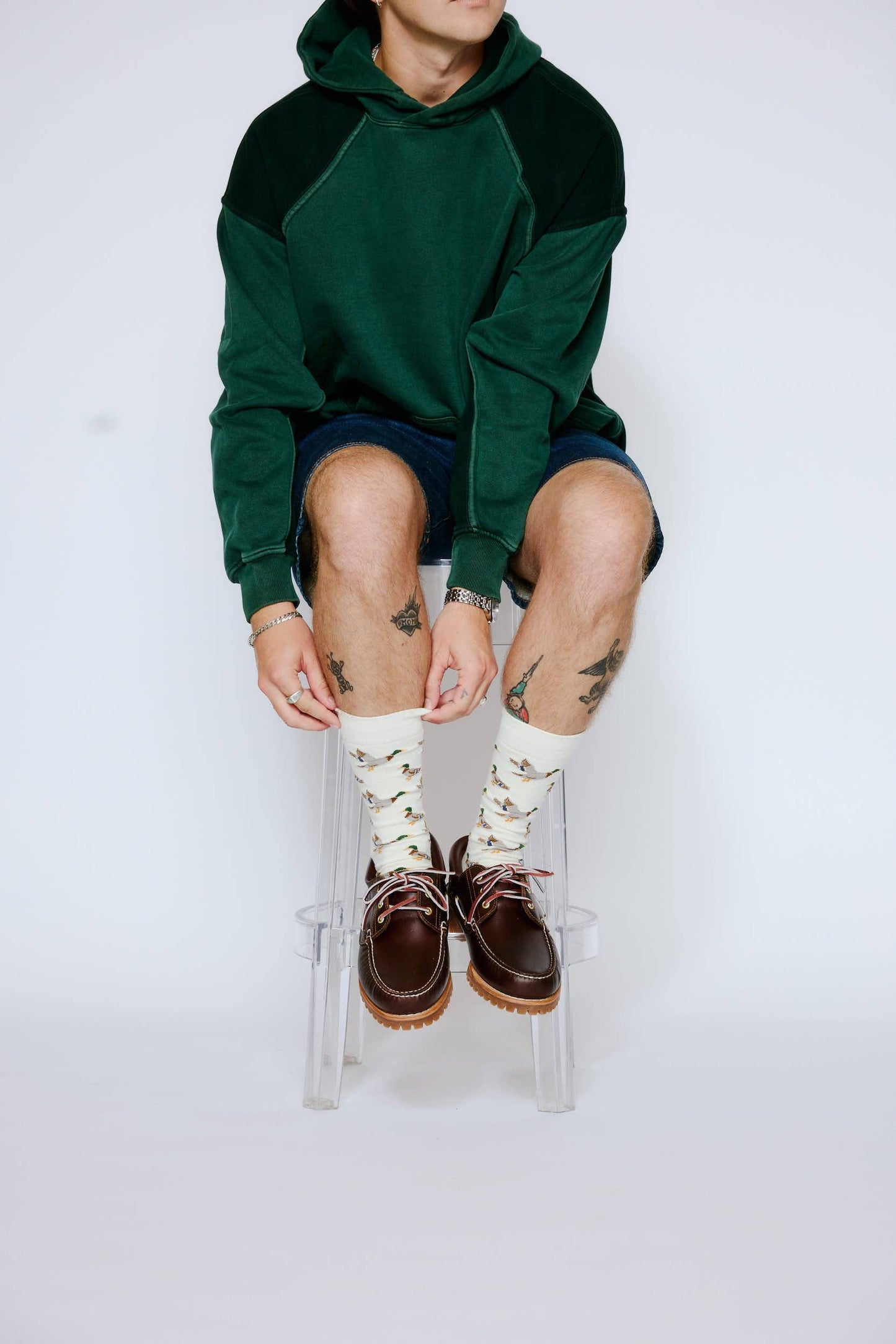 Person wearing a green hoodie, white socks with patterns, and brown shoes sitting on a clear stool against a white background.