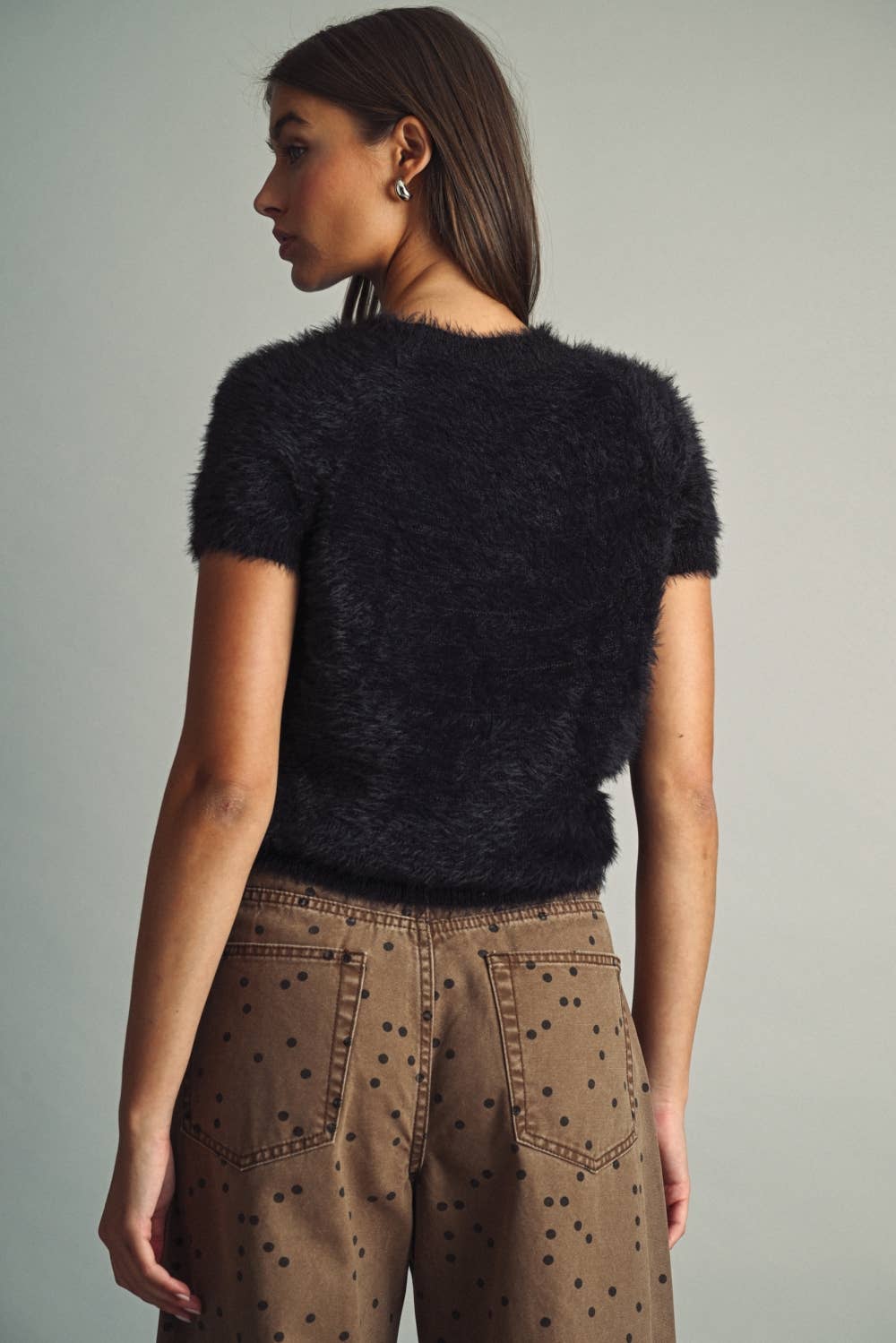 A woman shown from the back wearing a short-sleeve fuzzy black cropped top and brown pants with black polka dots, against a plain light gray studio background.