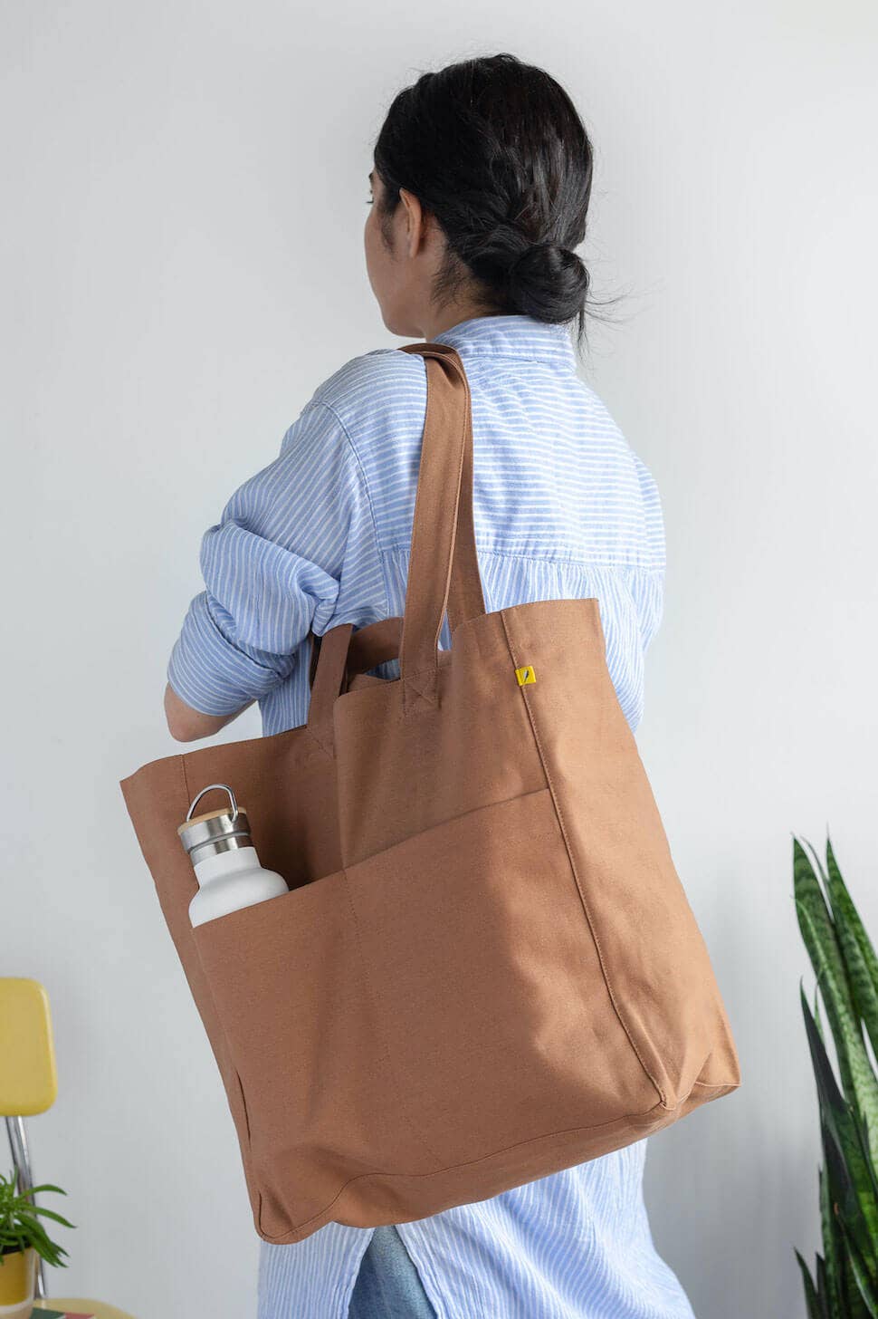 A woman wearing a light blue striped shirt is shown from behind, carrying a large brown canvas tote bag over her shoulder. The tote bag has a large front pocket holding a white bottle and features a small yellow tag.