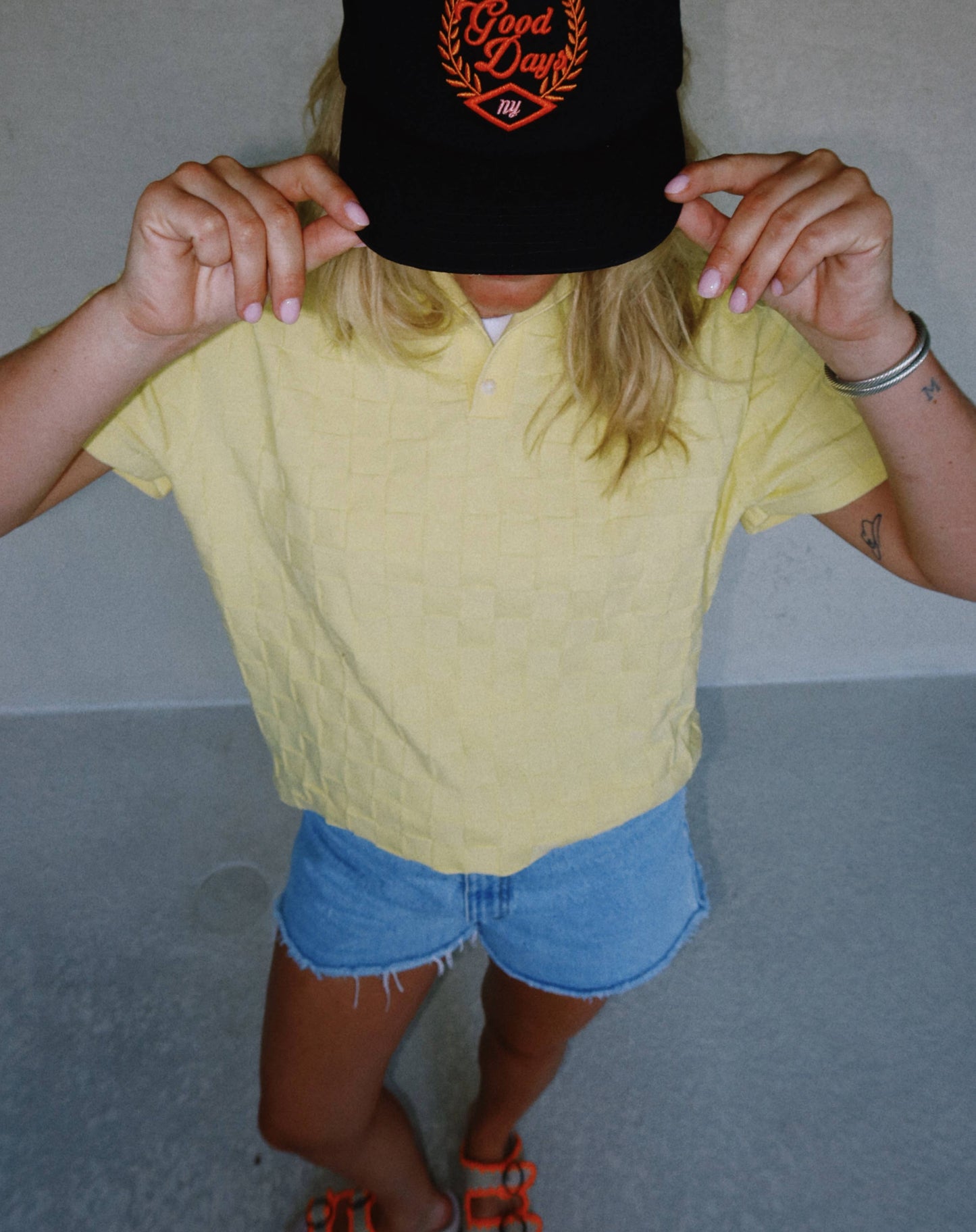 Person wearing a yellow shirt and denim shorts with a black cap featuring red text.
