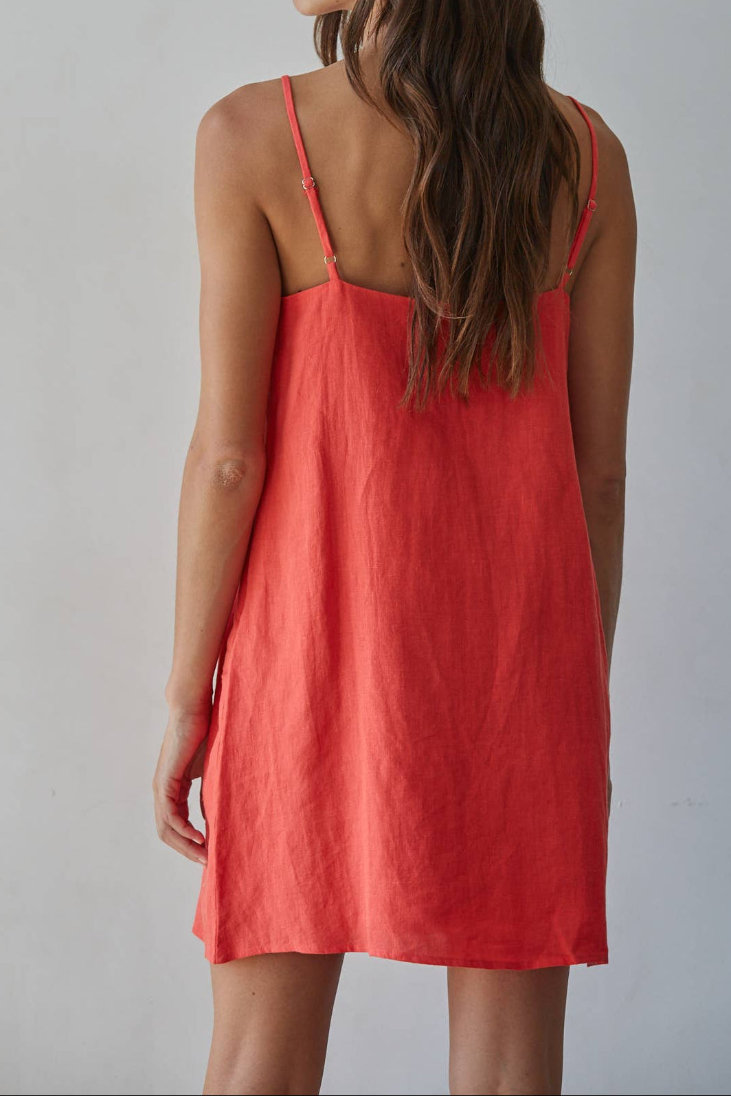 A woman wearing a coral red linen slip dress, shown from the back against a neutral light gray background.