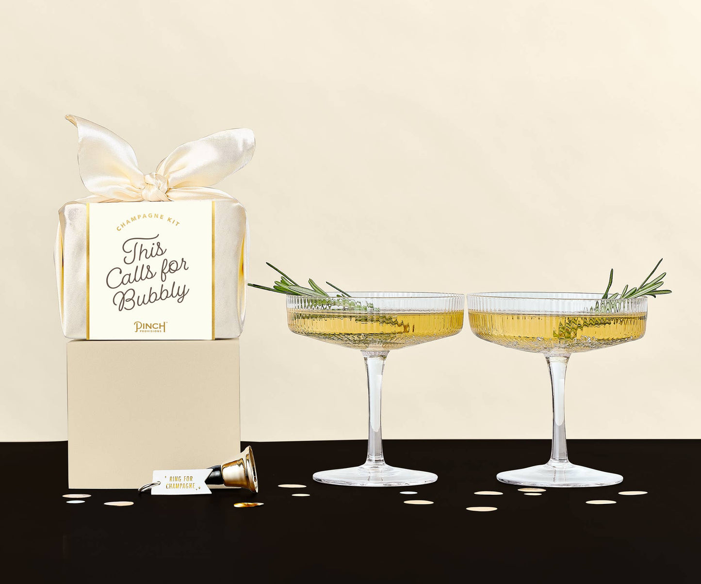 Two champagne glasses with decorative herbs and a gift box labeled 'This Calls for Bubbly' on a beige background.