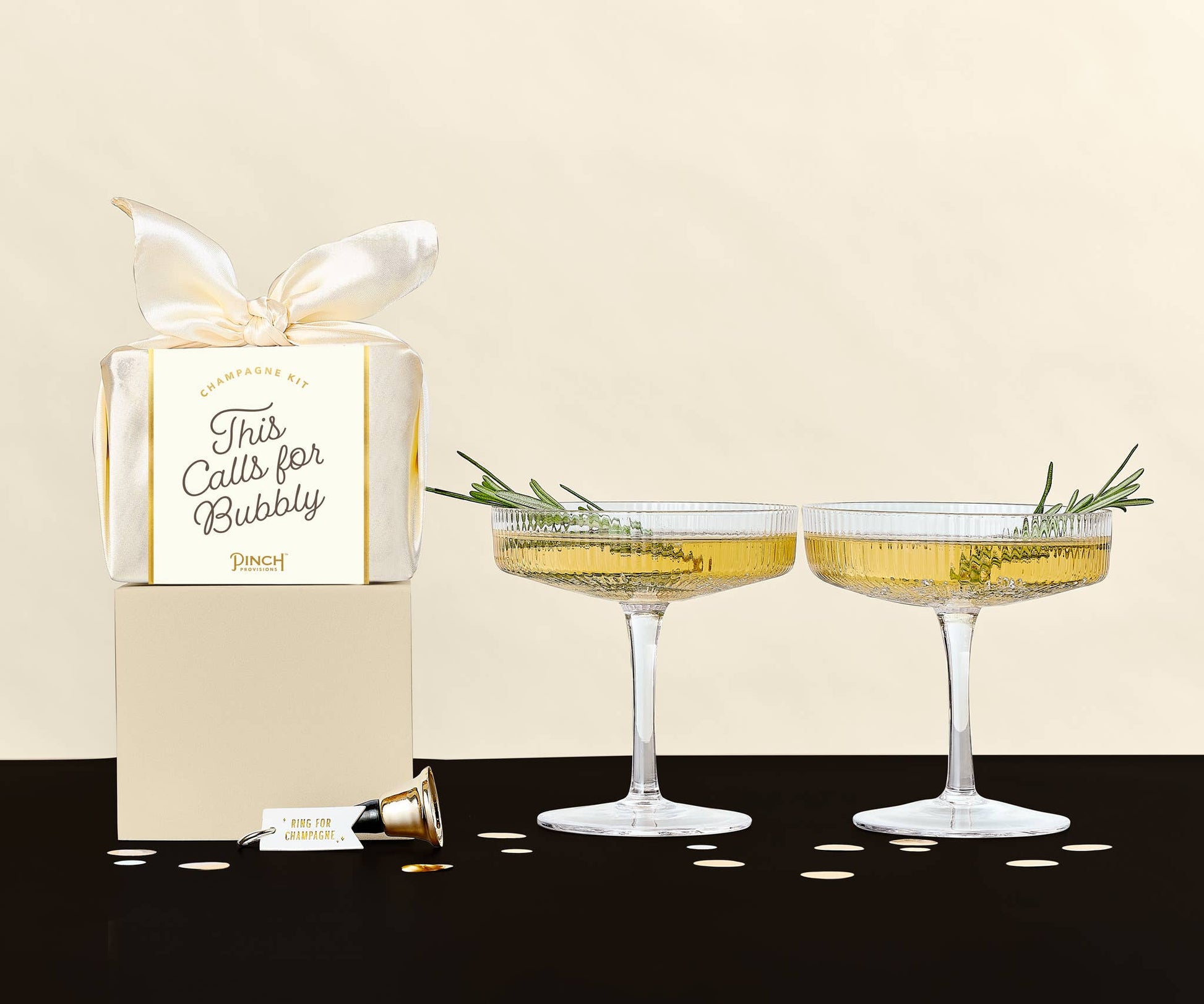 Two champagne glasses with decorative herbs and a gift box labeled 'This Calls for Bubbly' on a beige background.