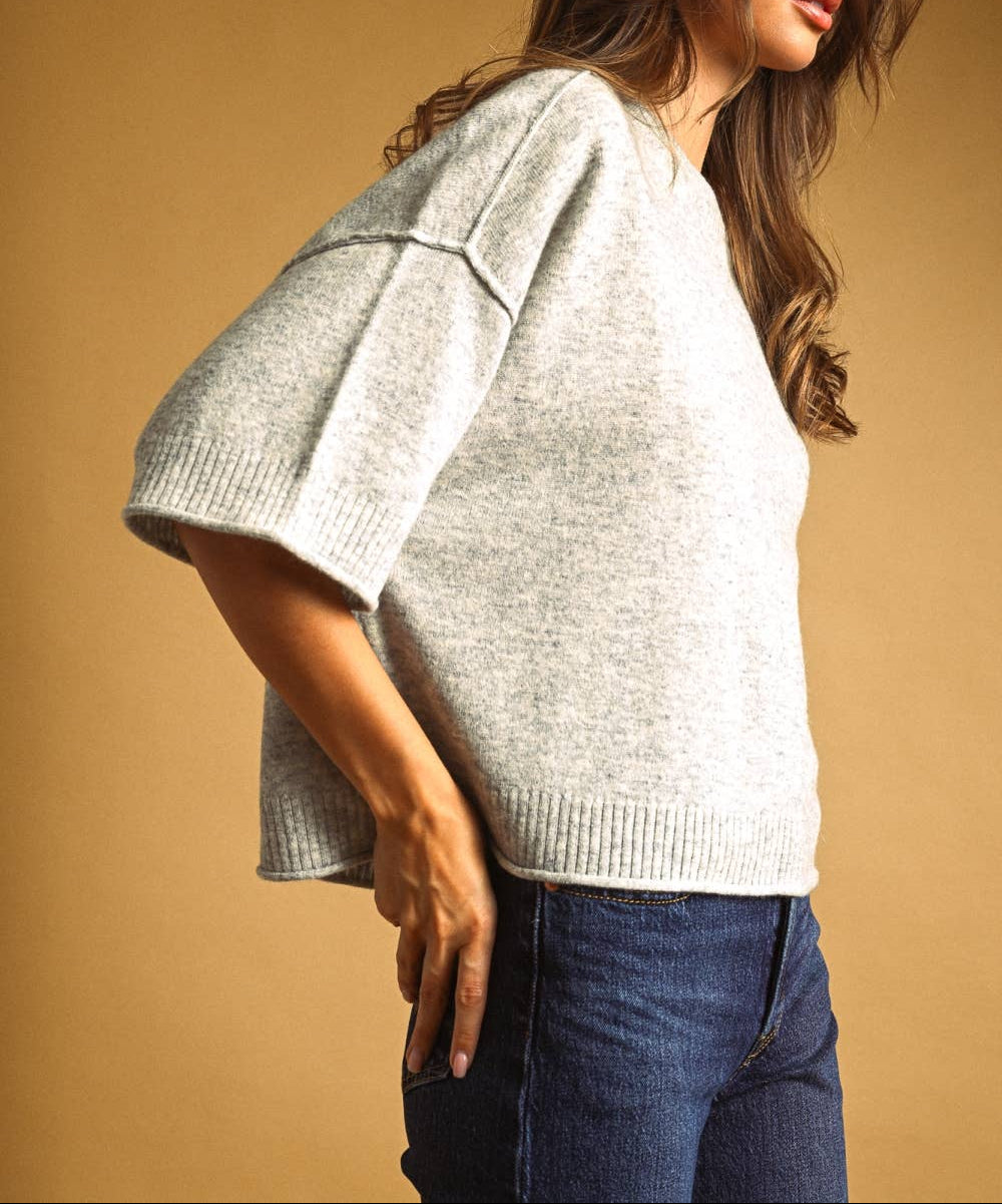 A woman wearing a light gray heathered knit sweater with wide, short puff sleeves and a V-neck, paired with dark blue jeans, set against a solid warm tan background.