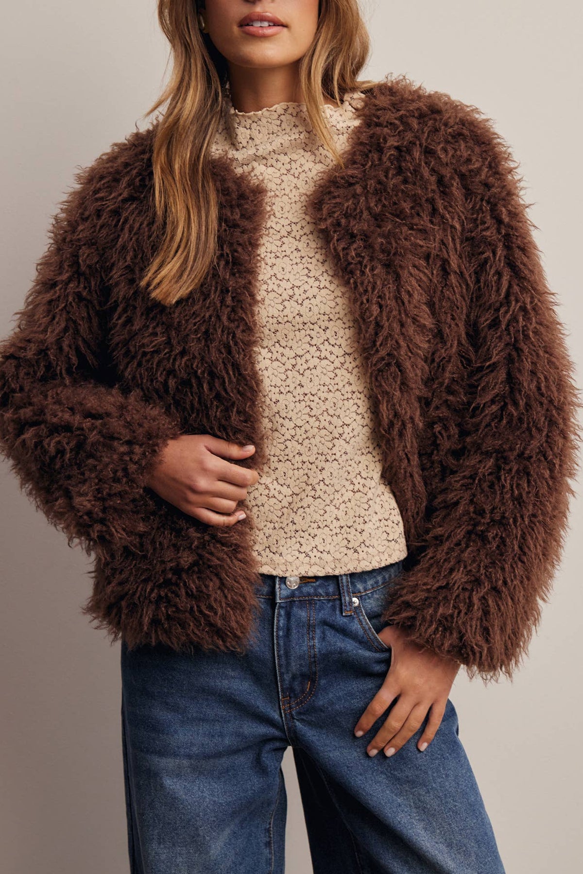 A woman wearing a brown shaggy faux-fur jacket over a beige knit sweater, blue jeans, and a brown New York Yankees baseball cap, posed against a plain light gray studio background.