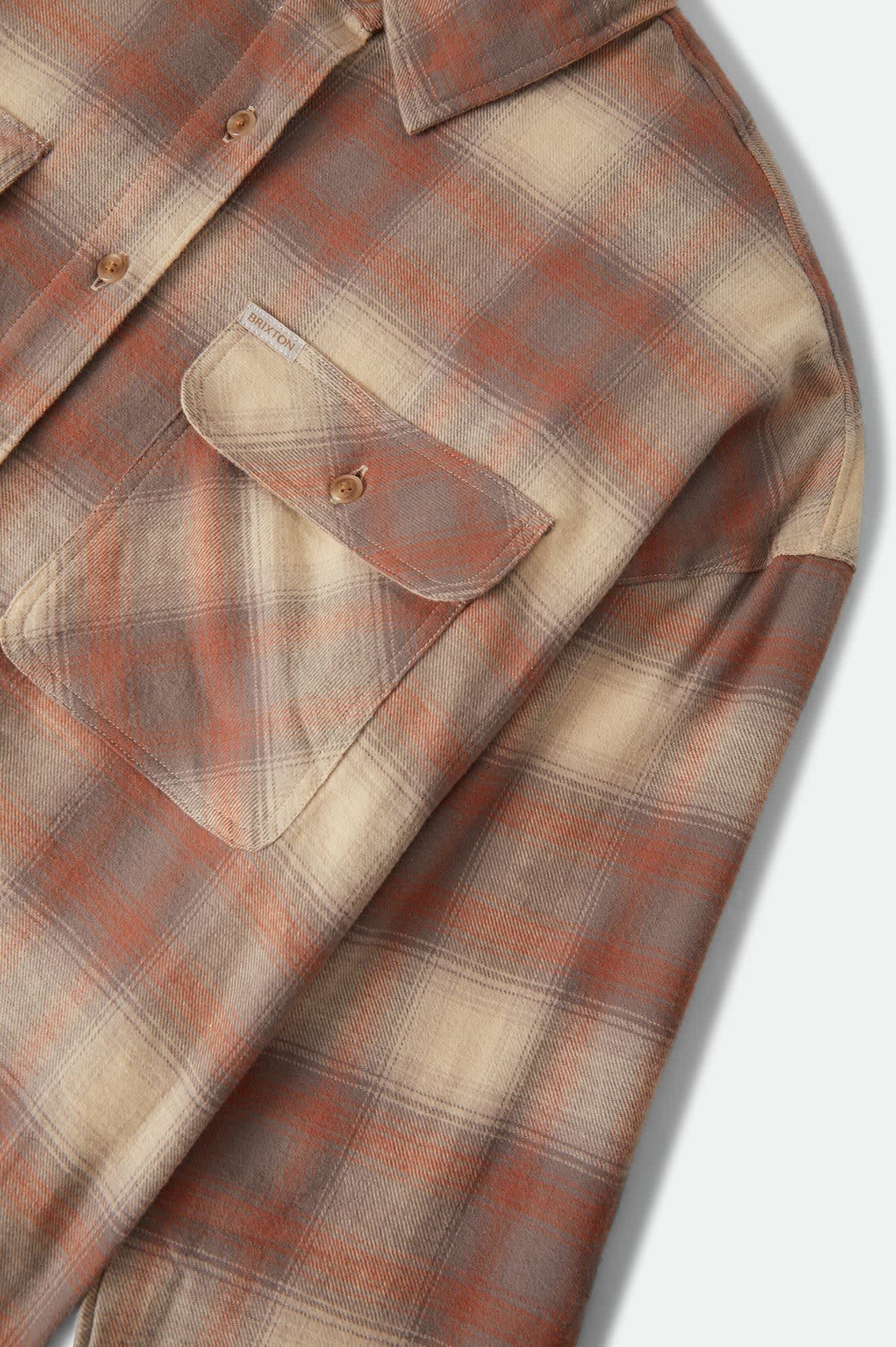 Close-up of a plaid button-up shirt in muted rust orange, beige, and gray tones, showing part of the chest pocket and collar.
