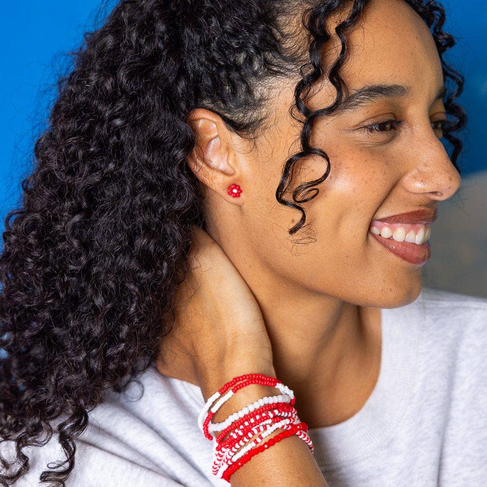 A close-up lifestyle shot of a smiling woman with curly dark hair, wearing a single small red stud earring and multiple red and white beaded bracelets on her wrist, set against a bright blue background.