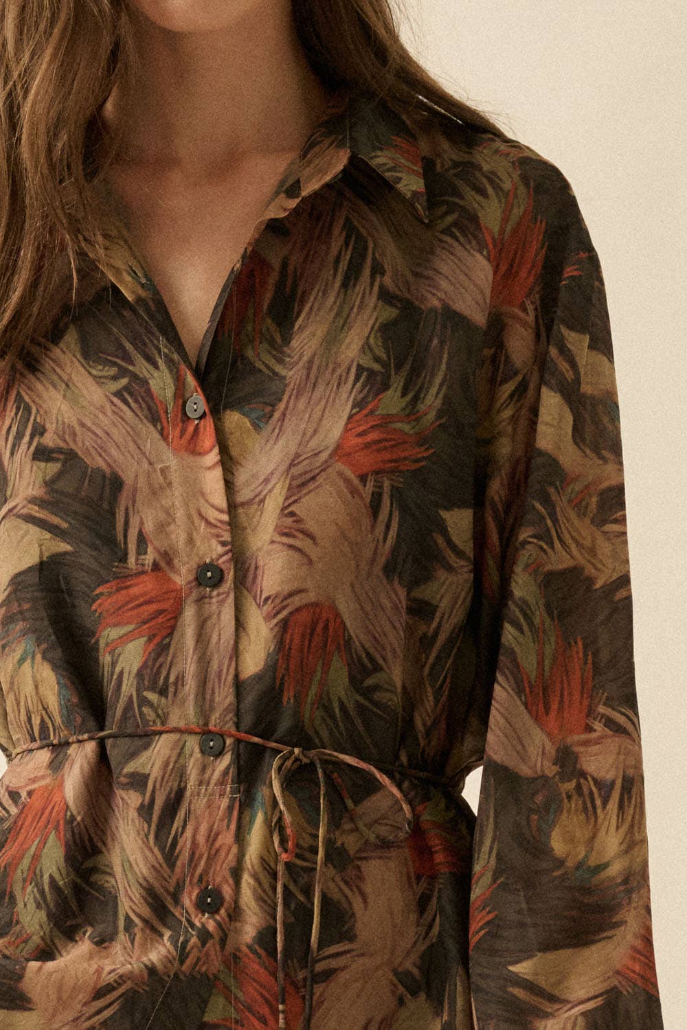 A close-up of a model wearing a long-sleeve button-front shirt dress with a tropical leaf print in earthy brown, beige, and rust-orange tones.