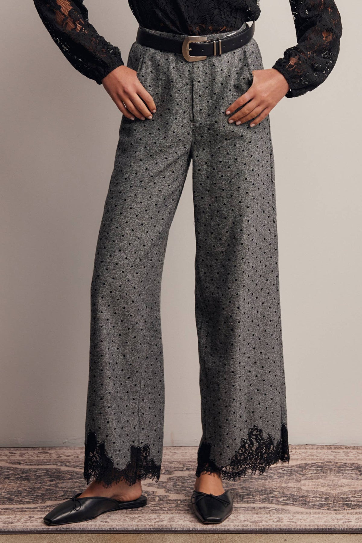 A person wearing high-waisted gray wide-leg trousers with a small black dot pattern, black lace trim at the hems, a black belt, and black flats, shown from the chest down against a plain studio background.