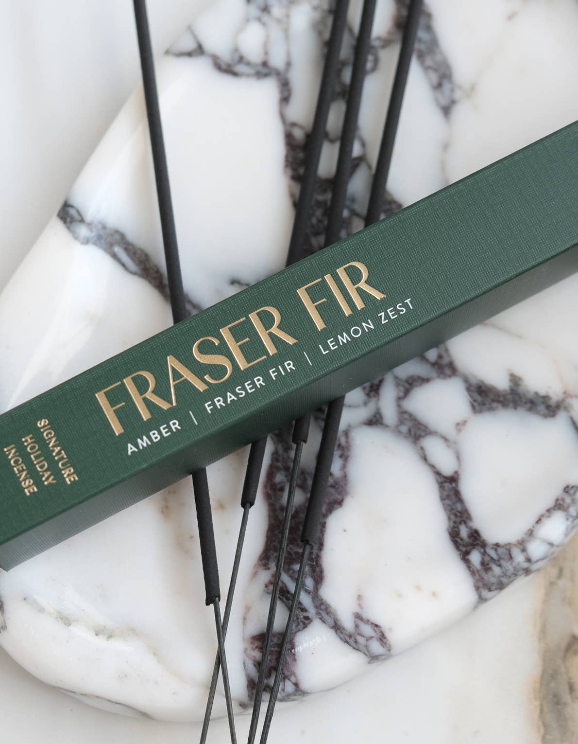 A dark green incense box labeled 'FRASER FIR' with gold lettering, placed diagonally over several thin black incense sticks on a white and gray marbled surface.