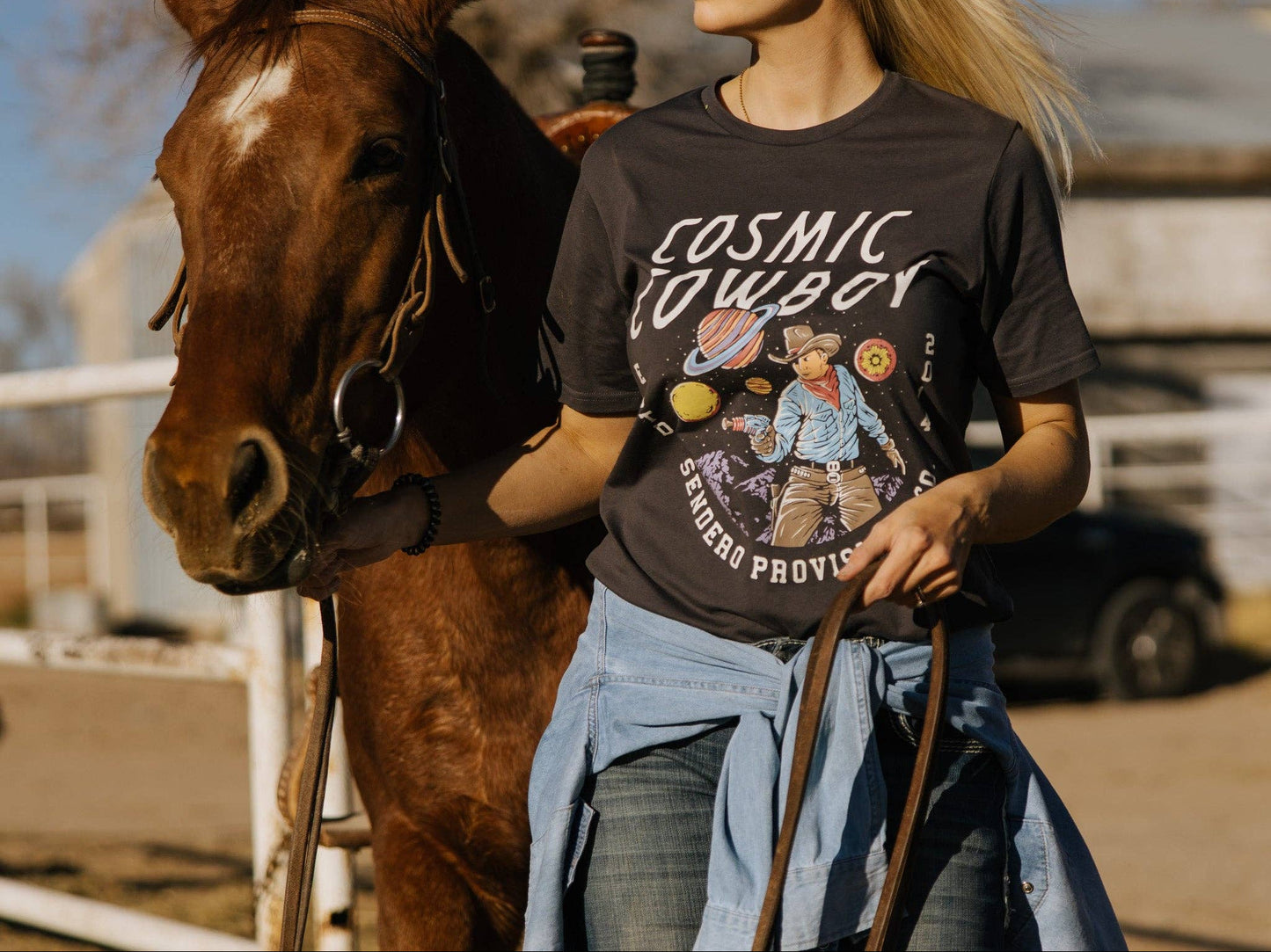 Woman wearing a 'Cosmic Cowboy' t-shirt standing next to a horse in an outdoor setting.