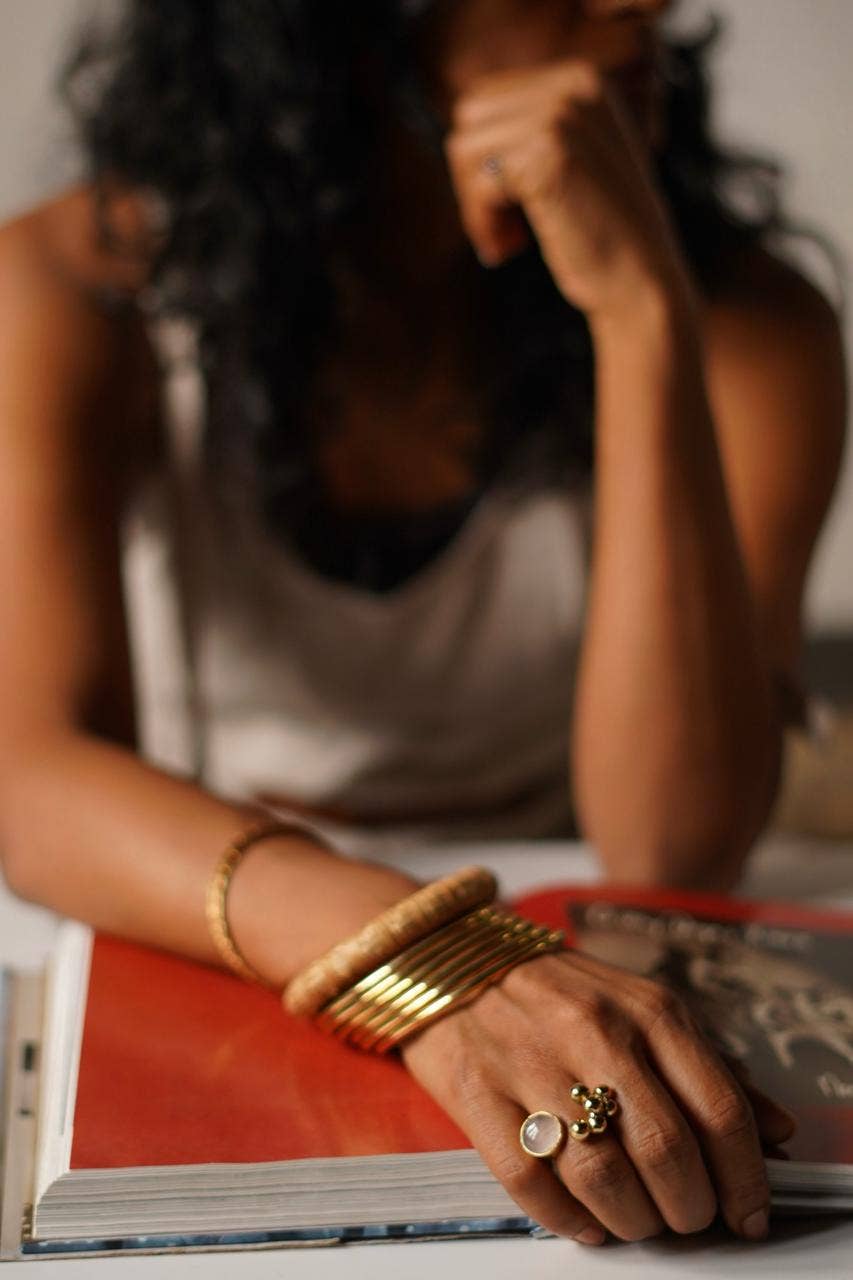 A close-up lifestyle shot of a person resting their head on their hand, wearing multiple gold bangles and rings while sitting at a desk with an open book.