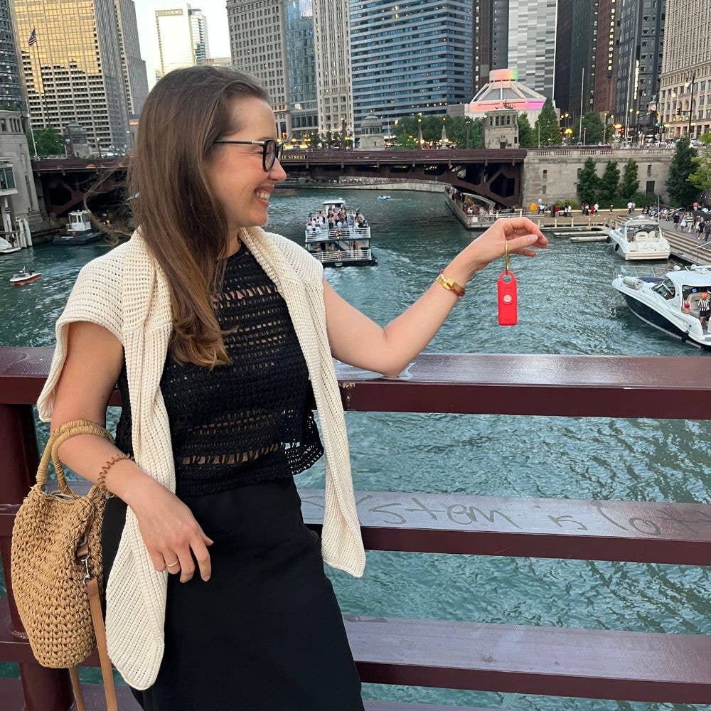 Woman holding a red keychain by a river with city skyline in the background
