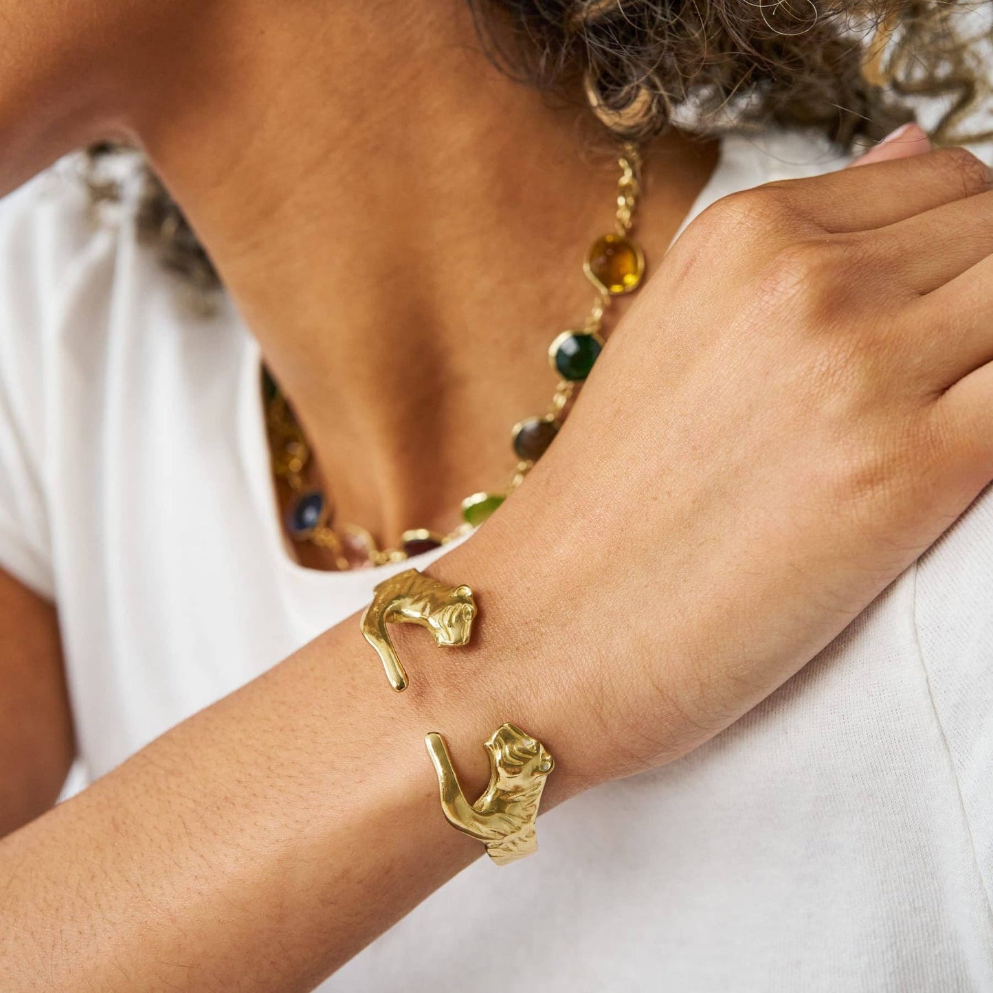 A close-up of a person's wrist wearing a gold cuff bracelet with two sculpted cat figures, paired with a white top and a beaded necklace.