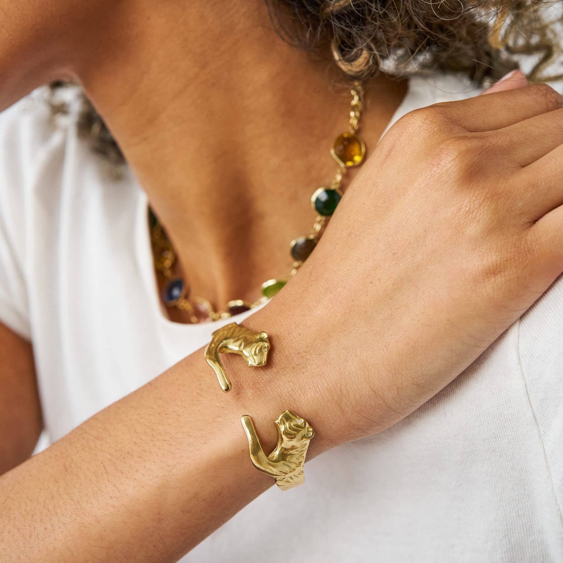A close-up of a person's wrist wearing a gold cuff bracelet with two sculpted cat figures, paired with a white top and a beaded necklace.