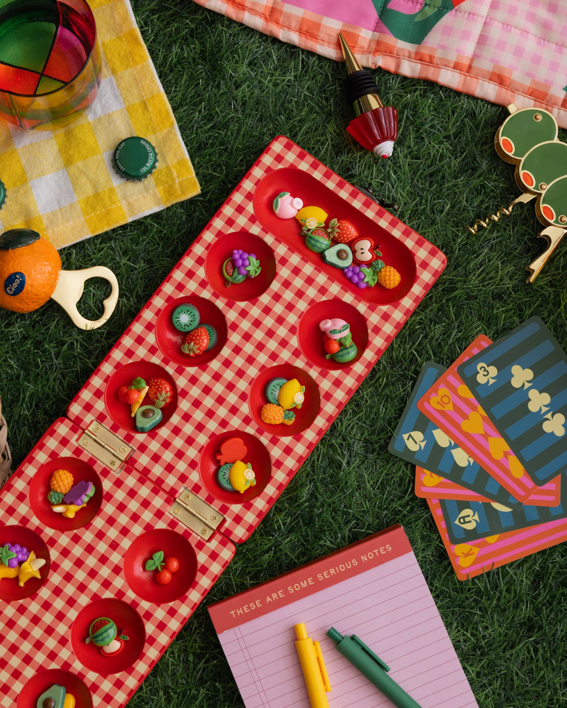 Colorful stationery items including a red checkered notebook, colorful cards, and a bottle opener on a grassy background.