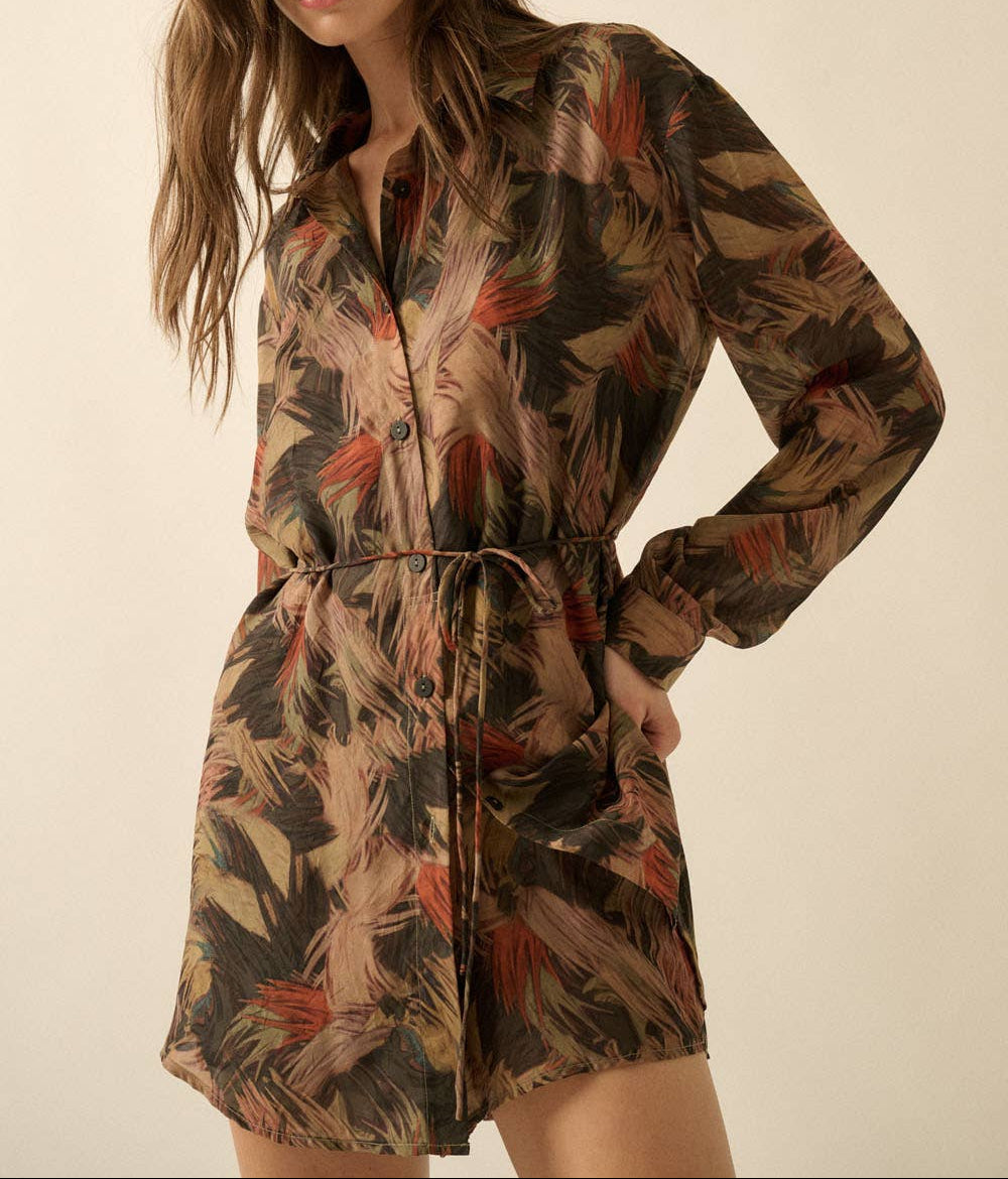 A woman modeling a long-sleeve mini shirt dress with a brown, beige, and rust-orange tropical leaf print against a plain beige studio background.