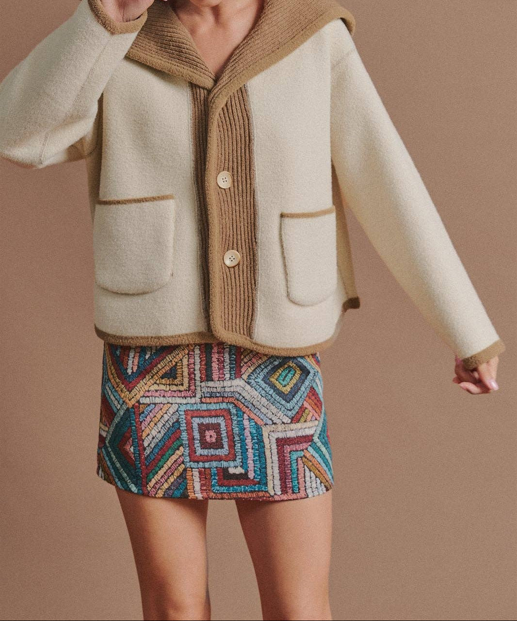 A woman models a cream-colored fuzzy jacket with a brown ribbed collar, cuffs, and button placket, paired with a colorful geometric patterned mini skirt and a brown cap, against a solid brown background.