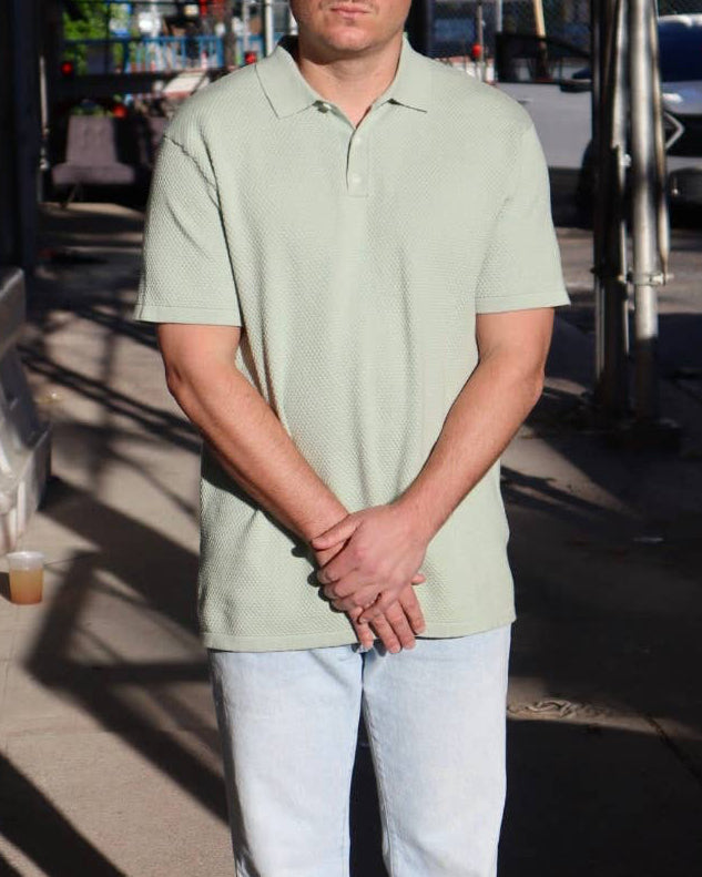 Man wearing a light green polo shirt and sunglasses standing on a city street.
