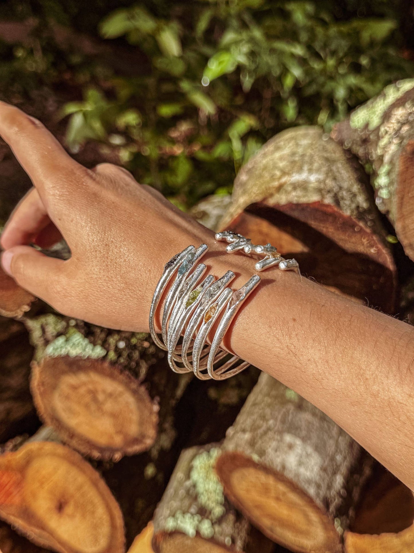 Hand wearing multiple silver bracelets with a natural background of logs and greenery
