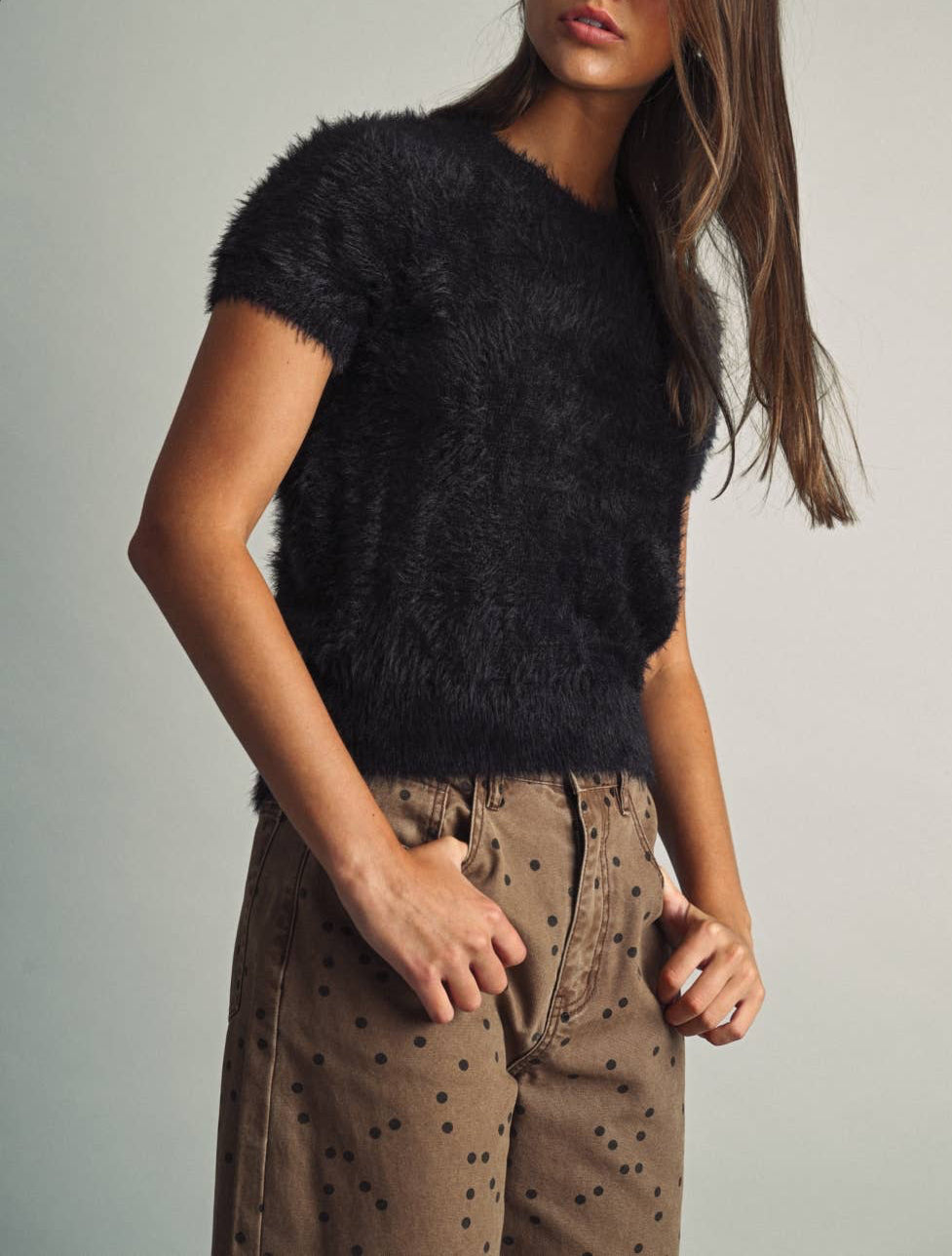 A woman wearing a black fuzzy short-sleeve knit top and brown pants with black polka dots, posing against a light gray studio background.