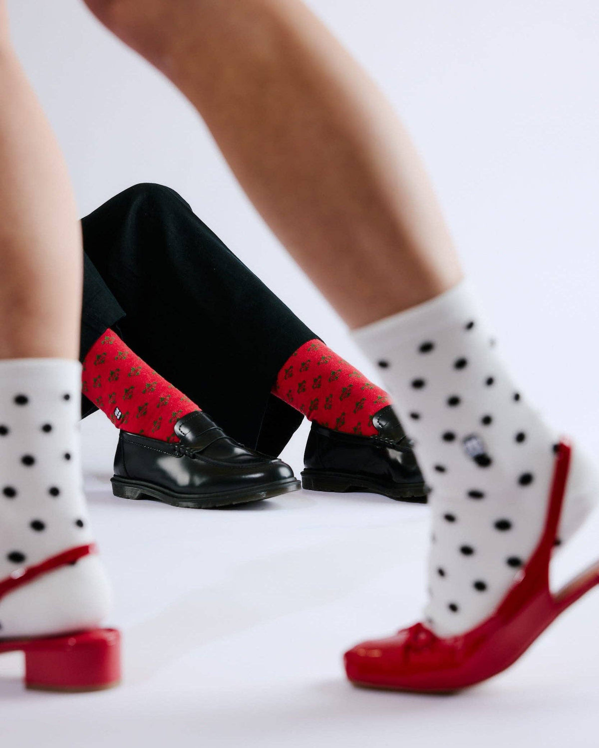 A close-up lifestyle shot of a person's legs and feet, showcasing three pairs of socks and shoes. The person is wearing black trousers, white socks with black polka dots, and red knit socks with a small repeating pattern, paired with red and black shoes.