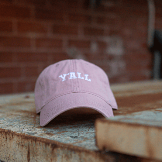 Pink cap with 'Y'ALL' text on a wooden surface with a brick wall background