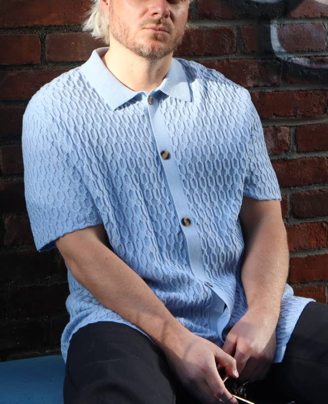 Person wearing a blue textured shirt and cap sitting against a brick wall.