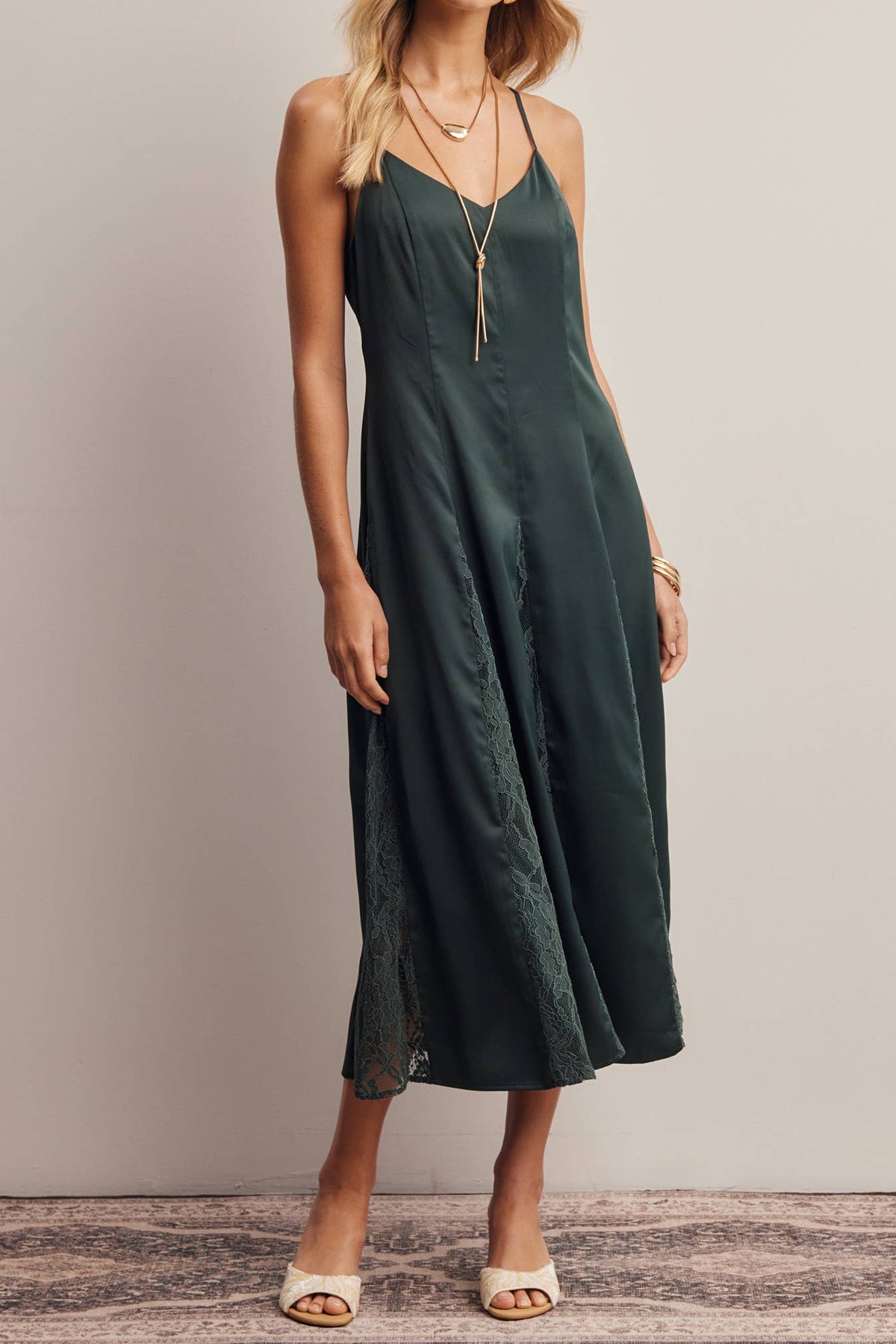 A woman modeling a dark forest green midi slip dress with thin spaghetti straps and a lace overlay at the hem, standing against a neutral beige wall.
