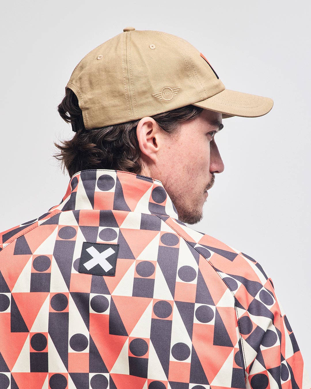Person wearing a beige cap and a colorful geometric-patterned jacket on a plain background