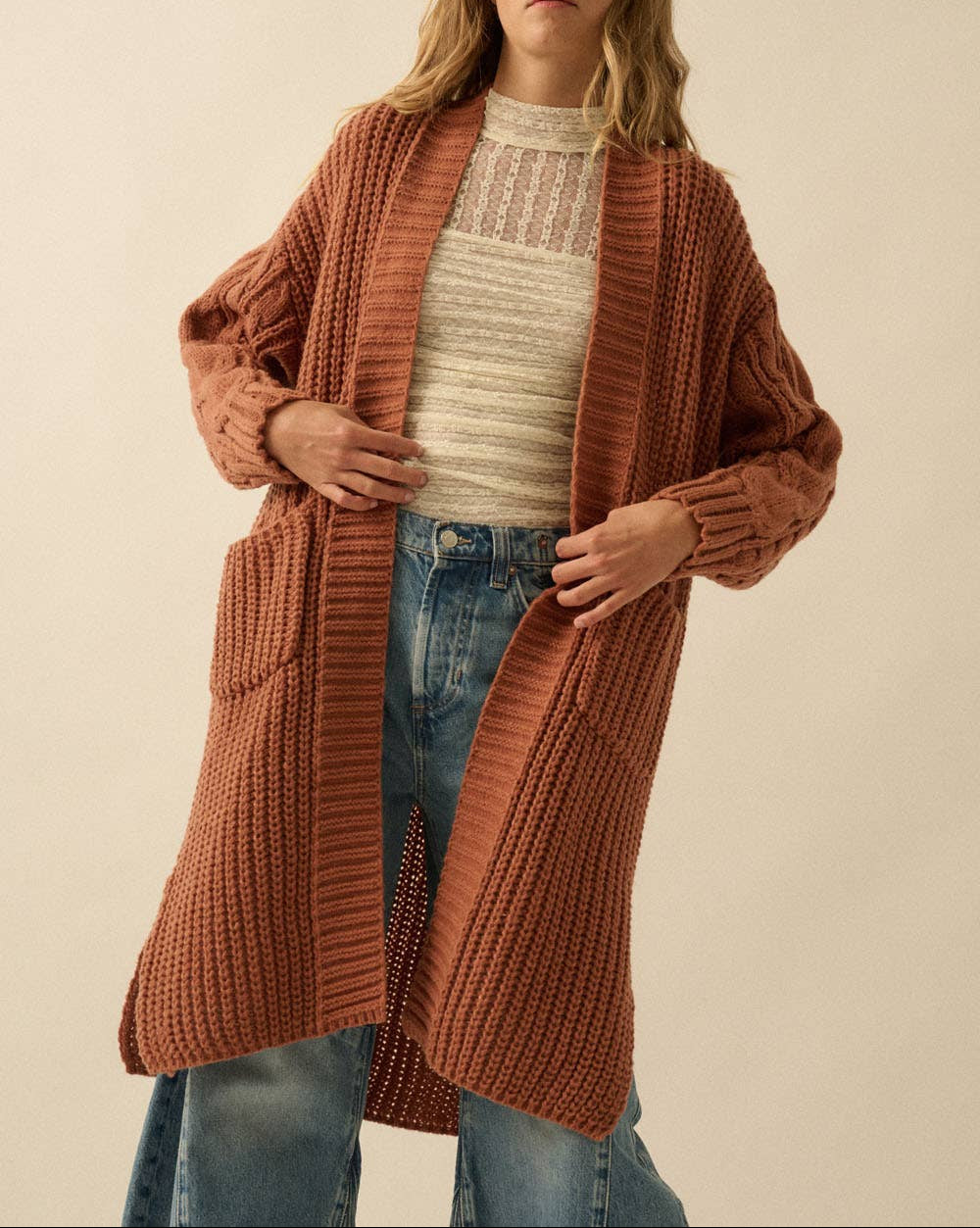 A woman wearing a long, rust-orange ribbed knit cardigan over a cream knit top and blue jeans, standing against a plain beige background.