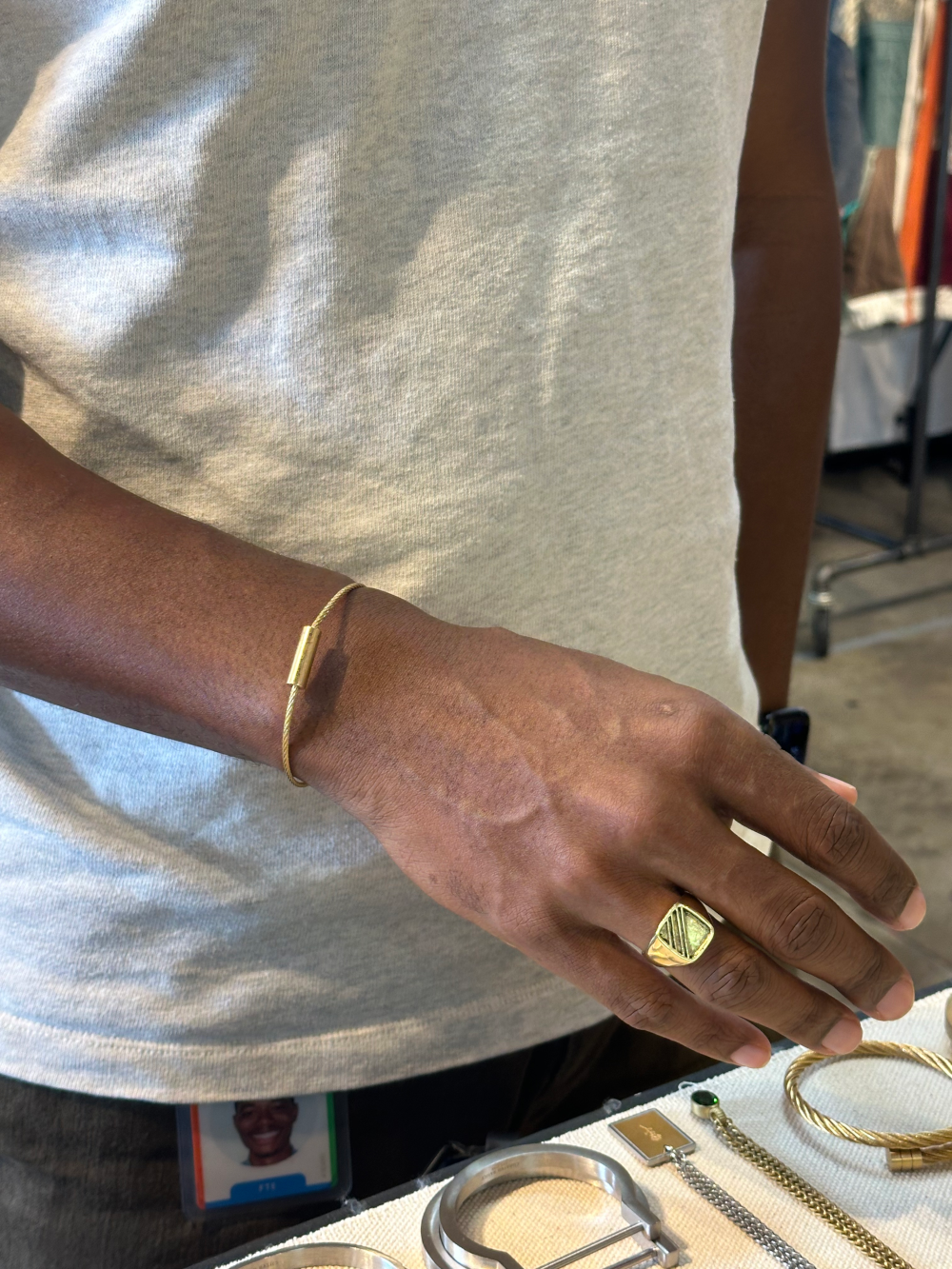 A close-up of a person's hand wearing a chunky gold signet ring and a slim gold bracelet, wearing a light gray t-shirt in what appears to be a retail or workshop setting.