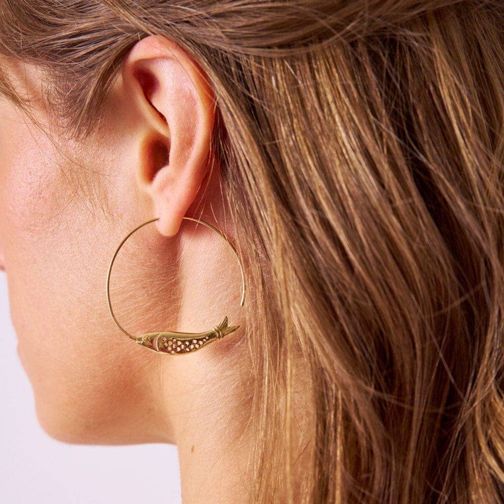 Close-up of a gold hoop earring with a small fish charm worn by a person.