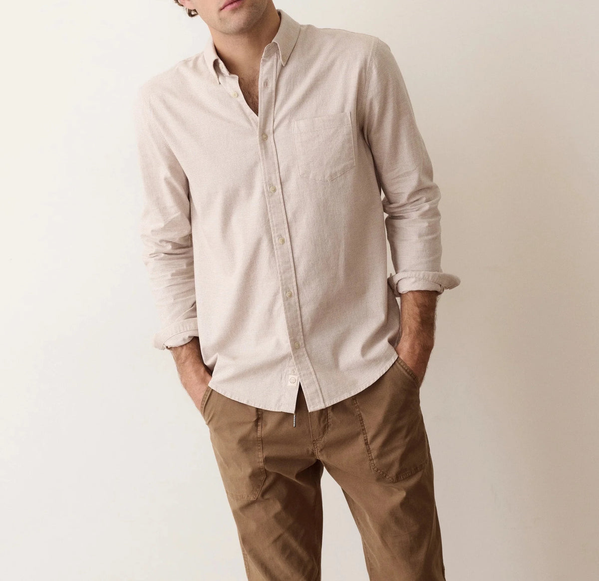 A man wearing a light beige linen long-sleeve button-down shirt and brown trousers, standing against a plain off-white background.