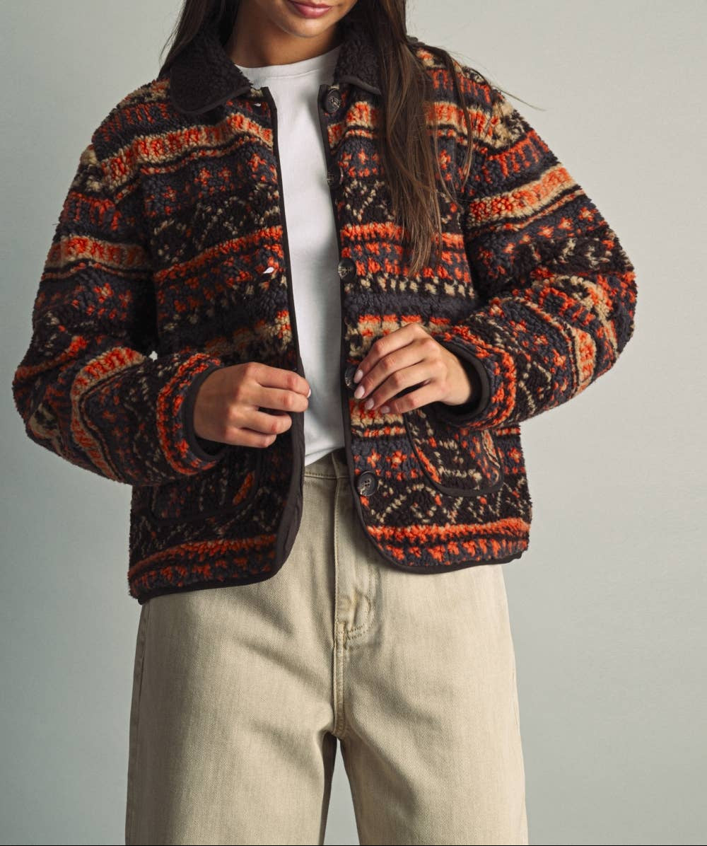 A woman wearing a cropped, patterned knit jacket with a dark brown collar and cuffs, featuring orange, beige, and black geometric designs. She is wearing a white t-shirt and beige pants, standing against a neutral gray background.
