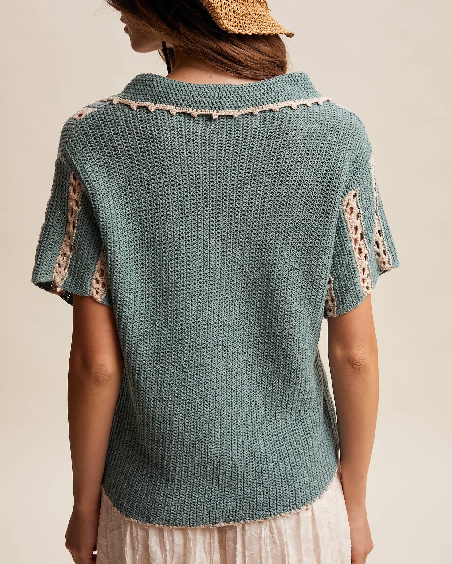 Woman wearing a teal knitted top with lace details and a straw hat on a neutral background