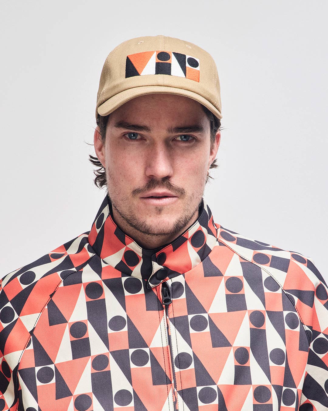 Man wearing a beige cap with geometric design and a patterned jacket on a plain background