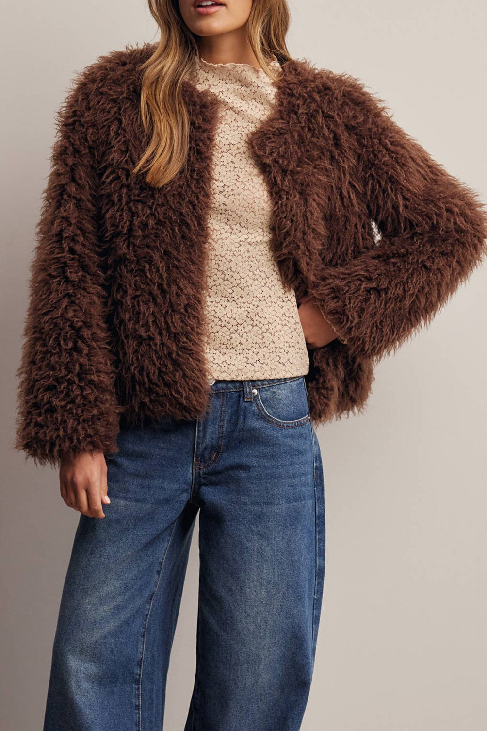 A woman modeling a brown shaggy faux-fur cropped jacket, a cream knit sweater, wide-leg blue jeans, and a brown baseball cap against a neutral studio backdrop.