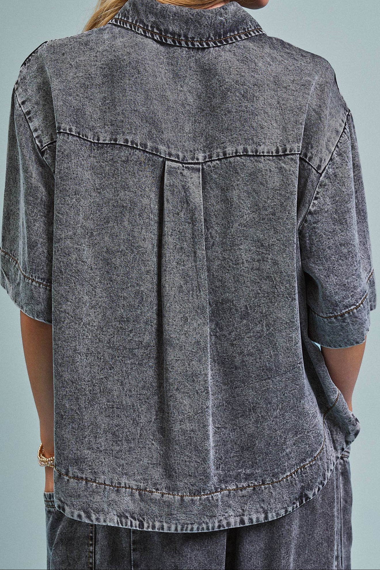A woman viewed from the back wearing a faded charcoal gray short-sleeve button-up shirt with a pleated back detail, paired with matching gray pants, against a solid light blue-gray background.