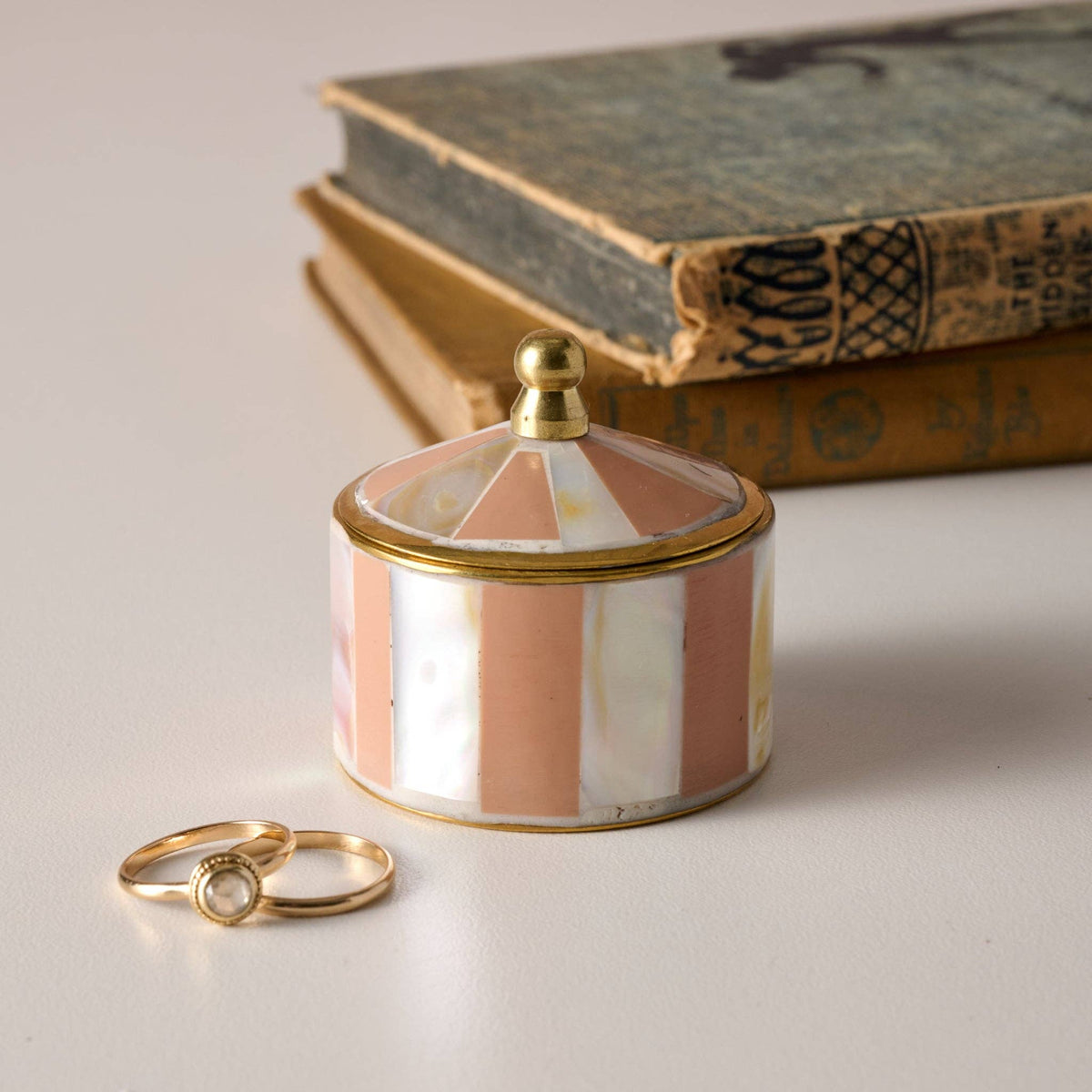 A small cylindrical jewelry box with a domed lid, featuring vertical pink and white stripes with gold trim and a gold knob, styled on a light surface with two gold rings and vintage books in the background.