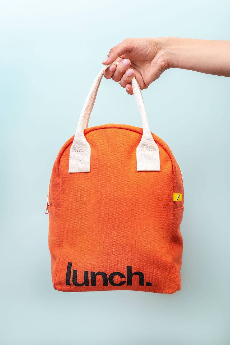 Orange lunch bag with 'lunch.' text held by a hand against a light blue background