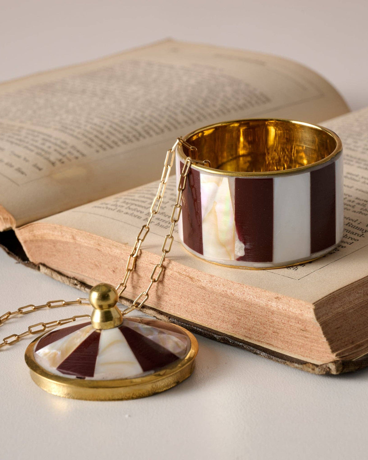 A gold-tone necklace with a round pendant and matching chain, displayed on an open vintage book next to a striped cuff bracelet. The jewelry features alternating dark red and white stripes with a glossy gold-tone finish.