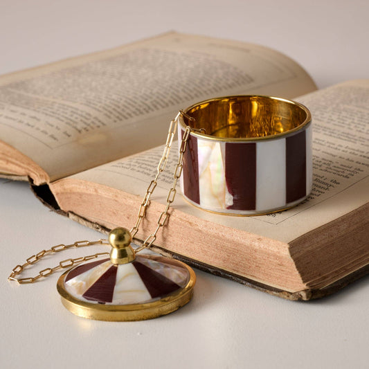 A gold-tone necklace with a round pendant and matching chain, displayed on an open vintage book next to a striped cuff bracelet. The jewelry features alternating dark red and white stripes with a glossy gold-tone finish.