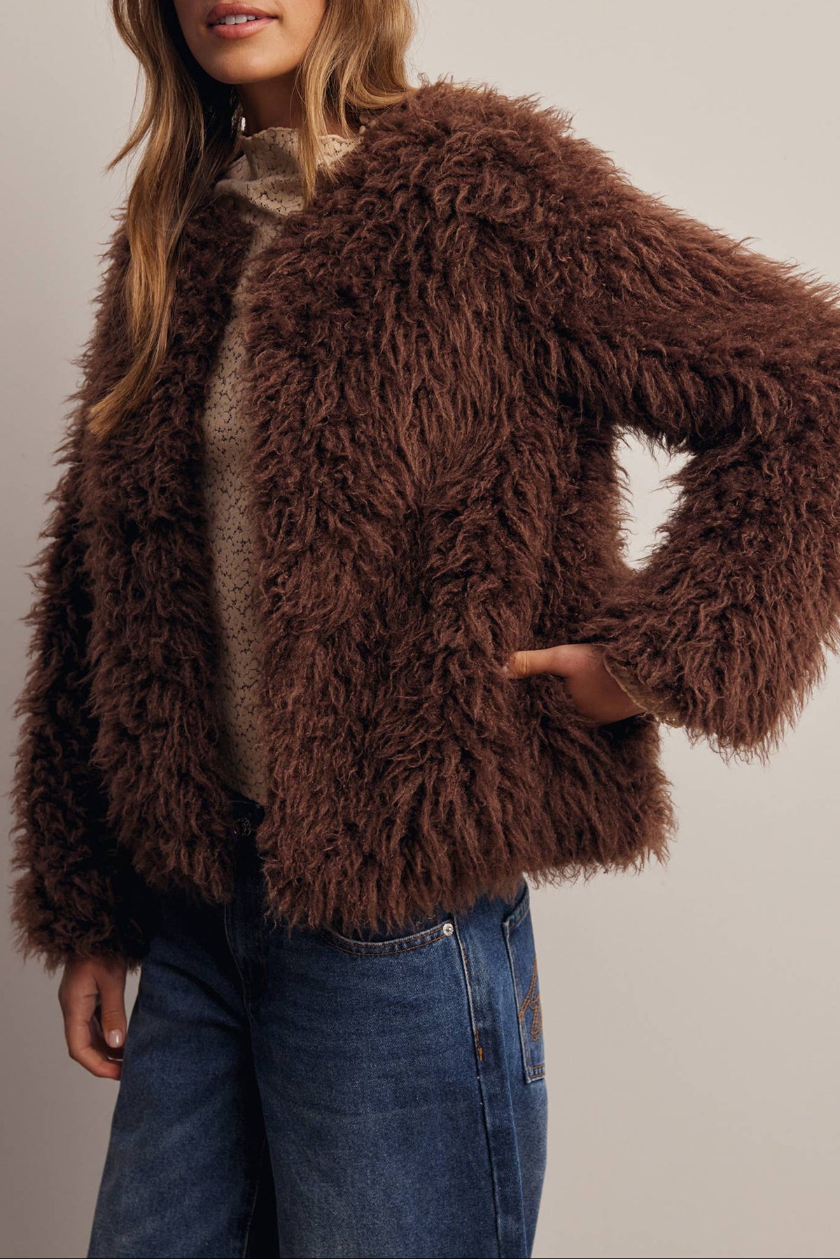 A woman wearing a brown shaggy faux-fur jacket over a beige top and blue jeans, with a brown baseball cap, photographed against a plain light gray studio background.