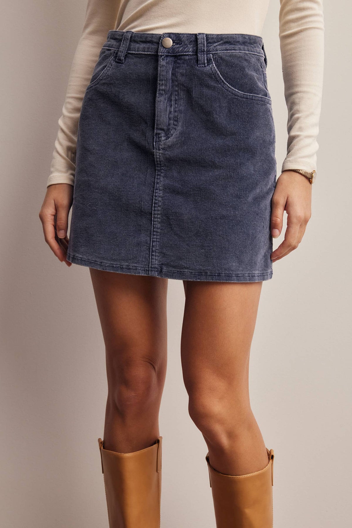 A model wearing a dark blue denim mini skirt with a button fly, paired with a cream long-sleeve top and tan boots, shown from the waist down against a solid beige background.