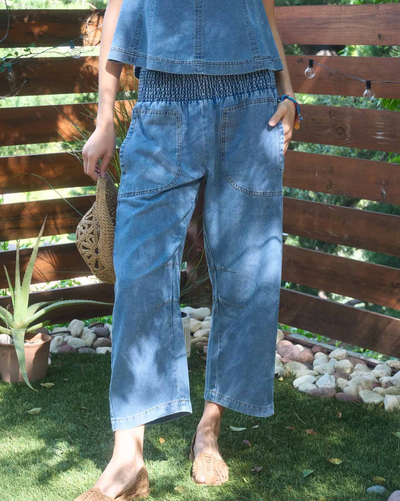 Person wearing a blue denim outfit standing outdoors with a wooden fence and plants in the background.