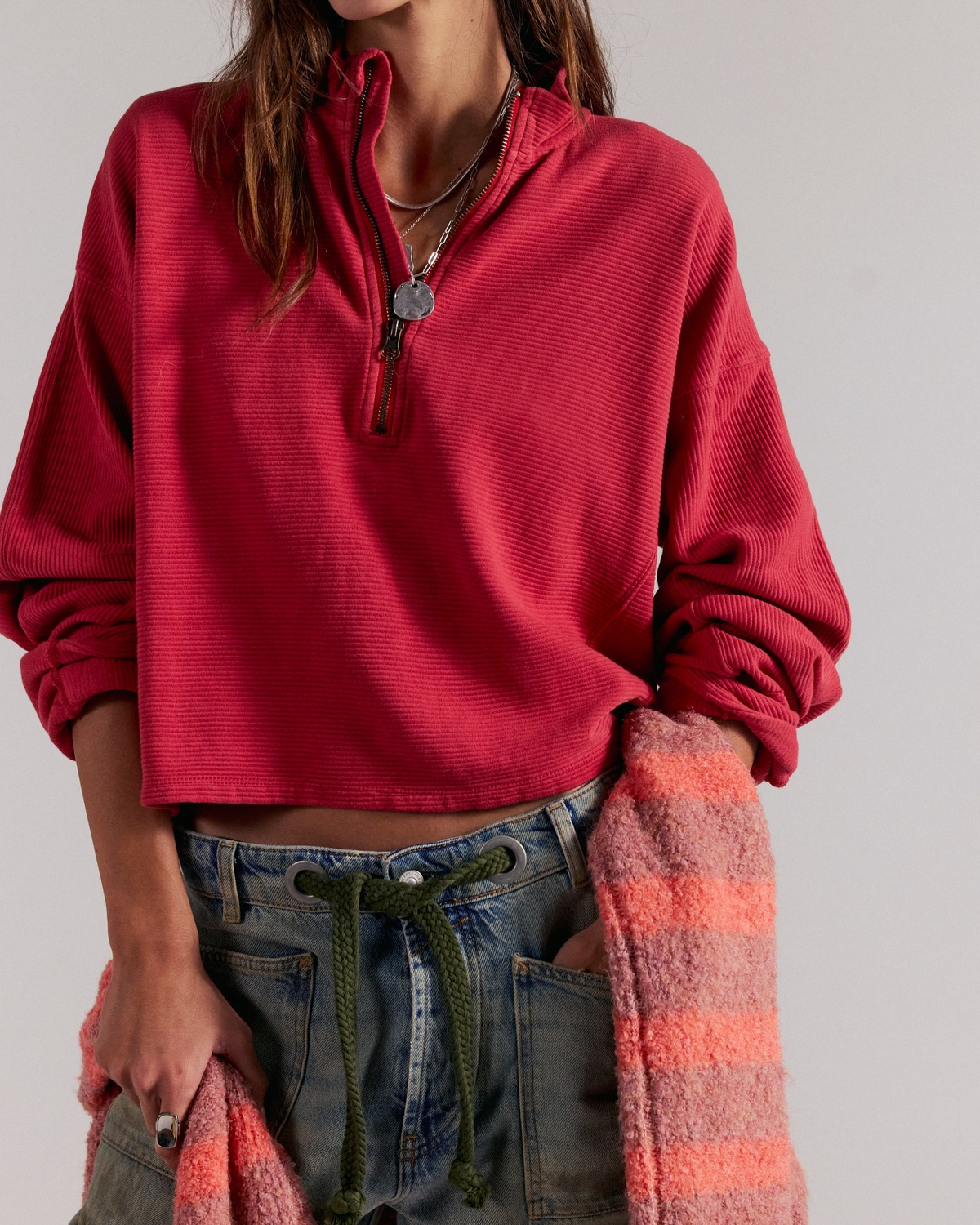 A person wearing a bright red ribbed quarter-zip pullover top with rolled sleeves, paired with blue jeans and holding a pink striped knit sweater against a plain light gray background.