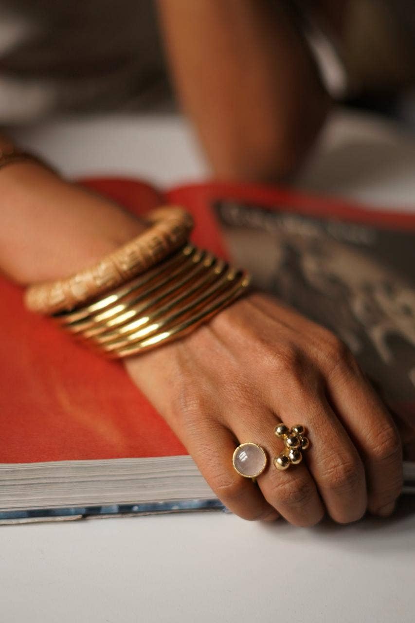 A close-up of a person's hand resting on an open book, showcasing gold jewelry. The hand is adorned with a stack of chunky gold bangles on one wrist and a statement ring featuring a large oval white stone and a small gold floral cluster on the ring finger.