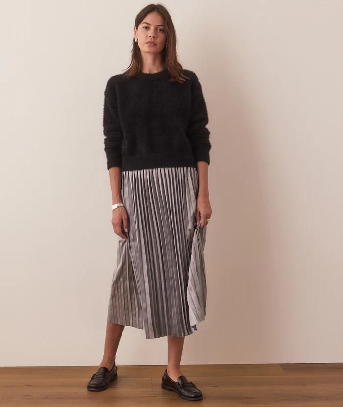 A woman standing wearing a black sweater and a midi pleated skirt with horizontal stripes. She is also wearing a watch and flat shoes.